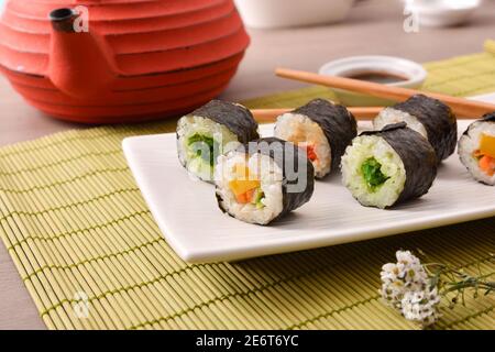 Detail of assorted sushi maki on white plate on wooden table with bamboo tablecloth. Front view. Horizontal composition. Stock Photo