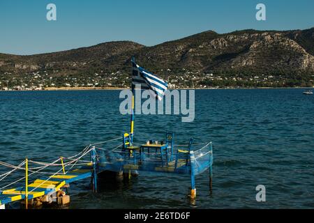 View of a Greek flag waves at a Wooden platform over the sea water leading to a table with three chairs for dinner during summertime next to the beach Stock Photo