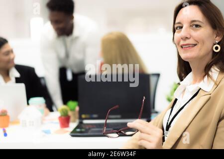 Portrait of young female teacher at desk looking to camera.   Employees working at computers in office with general manager. Job, study and Technology Stock Photo