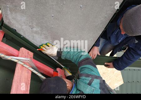 The worker on the roof of the house sheathes the chimney with a metal profile, the profile is green, the master uses a drill and is dressed in winter Stock Photo