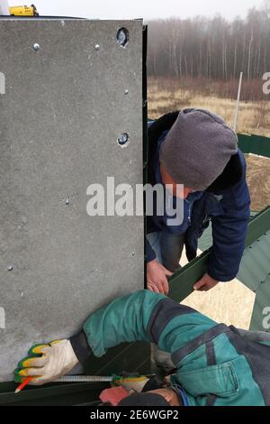 The worker on the roof of the house sheathes the chimney with a metal profile, the profile is green, the master uses a drill and is dressed in winter Stock Photo