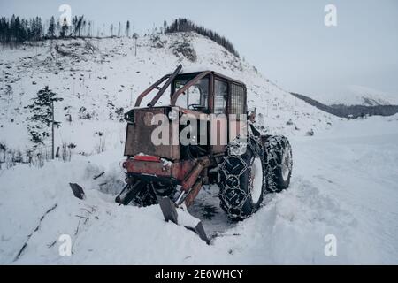 Winter scene in Europe, old tractor under the snow. Old Red farm tractor covered in fresh snow. Winter forest. Stock Photo