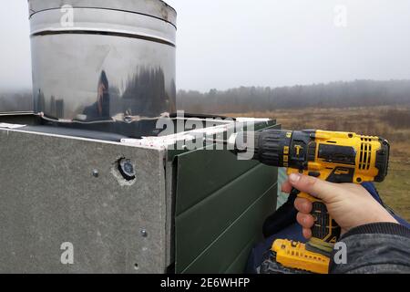 The worker on the roof of the house sheathes the chimney with a metal profile, the profile is green, the master uses a drill and is dressed in winter Stock Photo