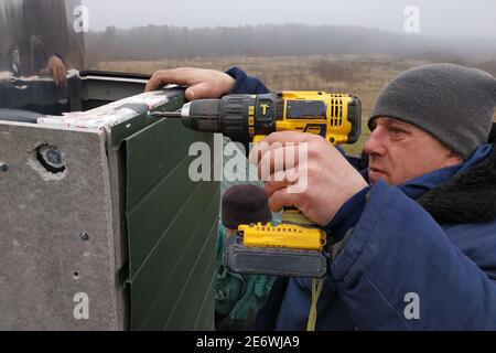 The worker on the roof of the house sheathes the chimney with a metal profile, the profile is green, the master uses a drill and is dressed in winter Stock Photo