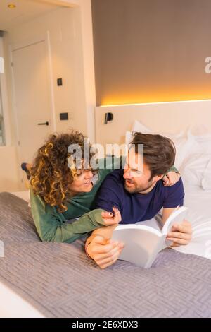 Vertical shot of a cute couple lying on a bed and reading an interesting book Stock Photo