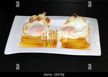 Two fried eggs with french fries (fried potatoes) cut into regular sticks placed on a rectangular plate and with a black background. Stock Photo