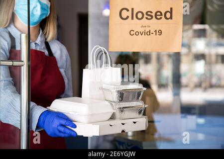 Woman waiter in protective medical mask and gloves work with takeaway orders. Waiter giving takeout meal while lockdown, coronavirus shutdown. Food Stock Photo