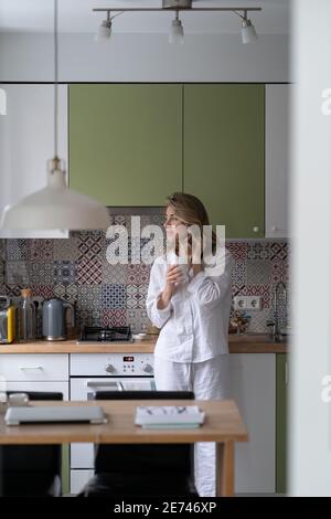 Pensive woman thinking, looking through the window, holding white mug, drinking tea at home Stock Photo