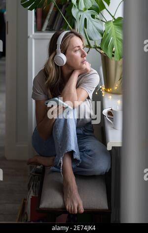 Woman listens to music wear wireless headphones using smart phone resting looking at window at home. Stock Photo