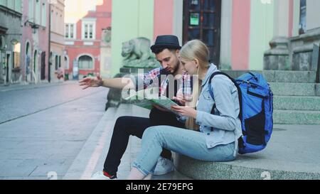 Happy tourist couple sitting on street stairs, checking city map and discussing. In the background there is beautiful old town view. Stock Photo