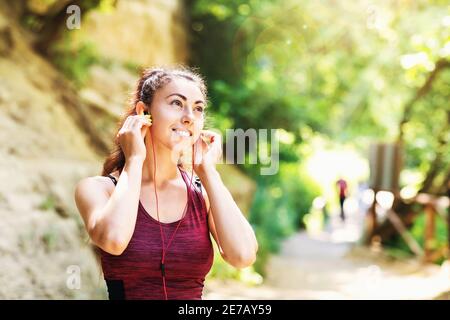 Young contented woman in sportswear in the park listens to music on headphones while exercising on the street. Healthy lifestyle concept.