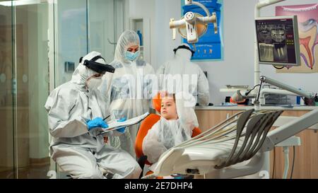 Pediatric dentist and nurse in ppe suit interrogating kid patient and taking notes on clipboard before examining. Stomatologist and assistent working in new normal dental office wearing coverall Stock Photo