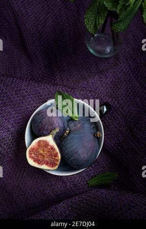 Overhead view of a cup full of delicious figs with some peppermint leaves on top of them next to a crystal jar. A purple cloth is in the background. Stock Photo