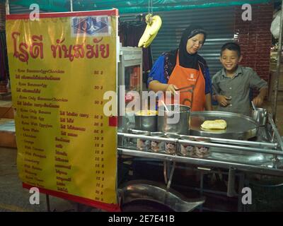 A young boy smiles happily as a mother who is a street food vendor teaches him how to make fried banana bread. The letters Thai mean their menu and pr