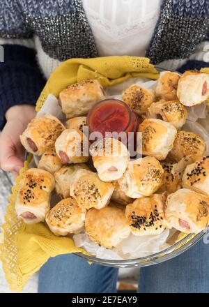 Dish full of homemade sausage rolls shown by a woman dressed in jeans. A jar of ketchup is in the middle. Stock Photo