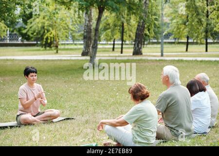Mature woman sitting on green grass snd talknig to senior people during therapy classes outdoors in the park Stock Photo