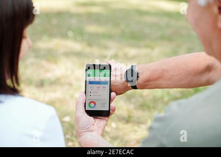 Rear view of woman showing sports app on her mobile phone and connecting it with smart watch Stock Photo