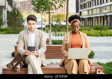 Two young business people sitting on bench and working online on computers outdoors Stock Photo