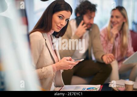 A young beautiful businesswoman wit a smartphone posing for a photo at a meeting in a relaxed atmosphere with her colleagues. People, business, meetin Stock Photo