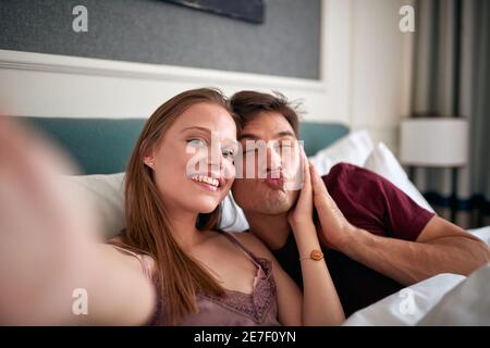 A young couple lying in the bed at the hotel room and taking funny selfies on a beautiful morning. Couple, love, hotel, together Stock Photo