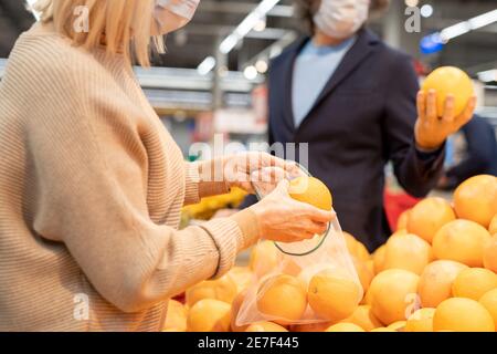 Mature couple in casualwear choosing fresh ripe oranges or grapefruits by display while woman putting one into fruit bag in supermarket Stock Photo