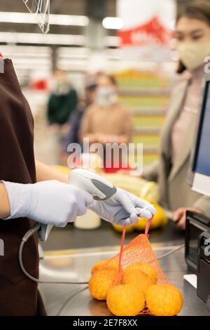 Gloved hands of young female cashier in uniform scanning pack of fresh oranges over counter by workplace while serving one of customers Stock Photo