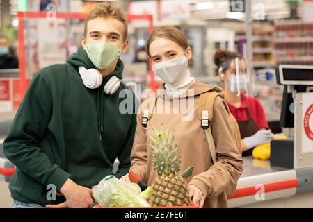 Happy teenage siblings in casualwear and protective masks standing in front of camera in supermarket against checkout counter and cashier Stock Photo