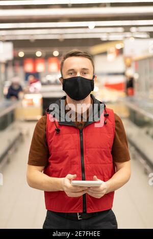 Young male courier in uniform and protective mask standing inside large supermarket and looking through online orders in touchpad Stock Photo