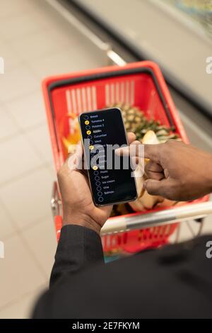 Hand of young African man with smartphone pointing at list of stuff to buy while pushing shopping cart with goods in modern supermarket Stock Photo