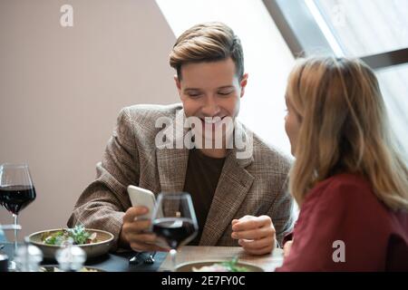 Happy young man in smart casualwear looking at screen of smartphone while watching curious video and showing it to his girlfriend Stock Photo