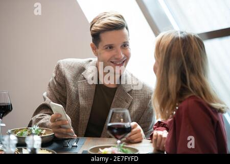 Happy young man with smartphone looking at his girlfriend with smile while talking to her by served table in luxurious restaurant Stock Photo
