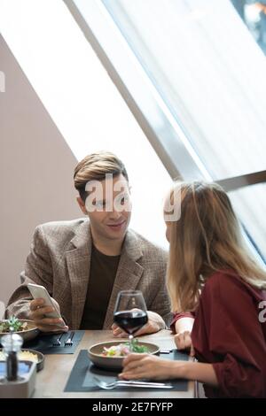 Elegant young man with smartphone looking at his girlfriend while discussing curious video with her by dinner in luxurious restaurant Stock Photo