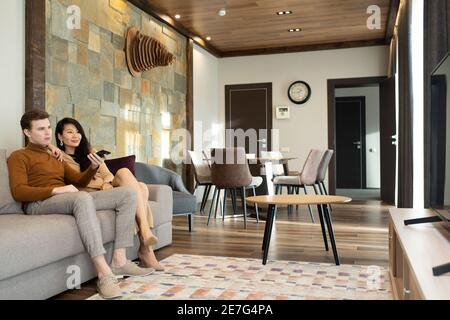 Young affectionate intercultural couple sitting on soft comfortable couch in hotel room and watching tv while Asian female using remote control Stock Photo