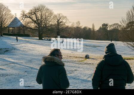 Two dads watch their children play in the snow in Golders Hill Park, London. The men wear winter clothing as the sun sets over Hampstead Heath, UK. Stock Photo