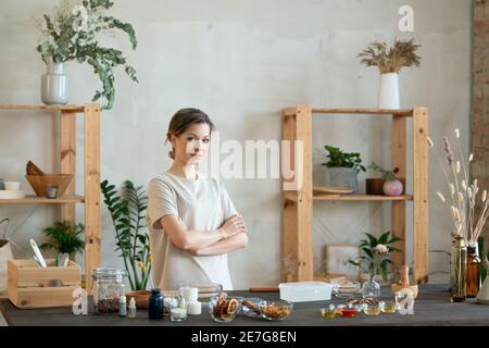Young confident woman in t-shirt crossing arms by chest while standing in front of table with ingredients for making natural cosmetic products Stock Photo
