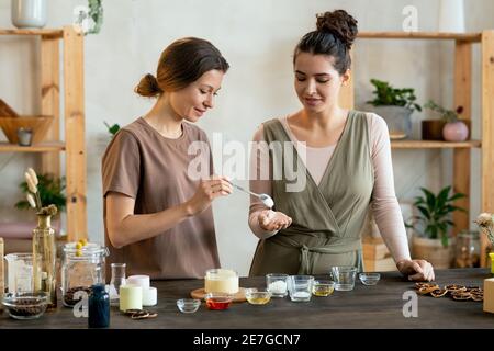 Young female putting blob of fresh handmade liquid soap mass on palm of her friend helping her with making natural handmade cosmetic products Stock Photo