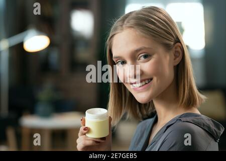 Young pretty female with blond hair holding small jar with natural handmade cosmetic product and looking at you with toothy smile Stock Photo
