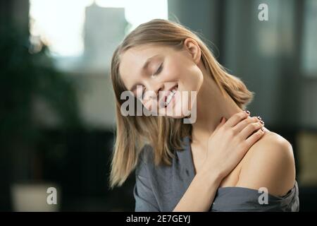 Young blond woman with toothy smile applying natural handmade cosmetic product on her neck and body and enjoying it in the morning Stock Photo