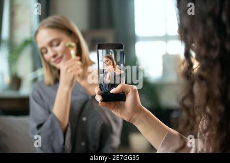 Hand of young woman with smartphone shooting her friend using massage roller upon her face and enjoying beauty procedure Stock Photo