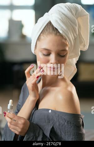 Young beautiful pleased woman in silk grey pajamas applying cosmetic product on her face and enjoying it in the morning after bath Stock Photo