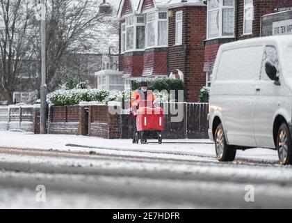Royal Mail British post man in deep cold snow falling on road and icy frozen pavement delivering letters trolley red uniform during severe weather Stock Photo