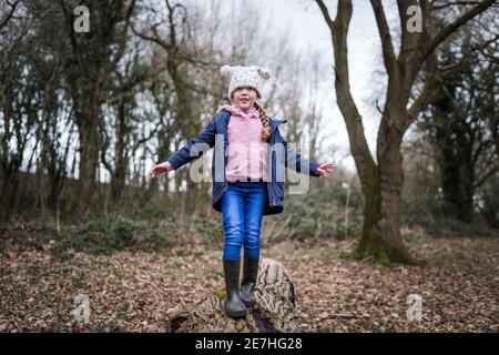 Excited happy young girl in wellinton boots sat in autumn countryside balancing on tree log with arms out exploring the great outdoors nature park Stock Photo