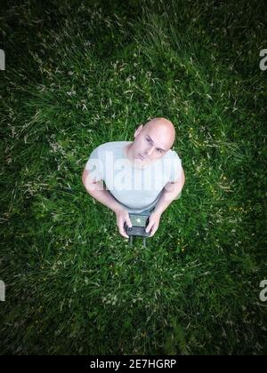Man alone looking up at flying drone from below on ground holding controller and mobile phone in hands stood in field look to sky wearing t-shirt Stock Photo