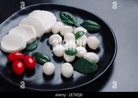 White small mozzarella cheese balls, spinach leaves and tomatoes on black plate. Stock Photo