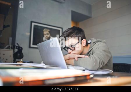 Tired boy with eyeglasses and a grey hoodie doing his homework with his notebook Stock Photo