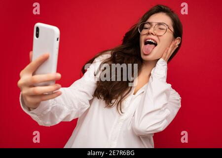 Closeup photo of beautiful positive young brunette woman wearing white shirt and optical glasses isolated over red background holding in hand and Stock Photo