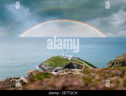 Colourful double rainbows in seascape over the ocean horizon with storm clouds dramatic sky and white lighthouse on top south stack island coastline Stock Photo