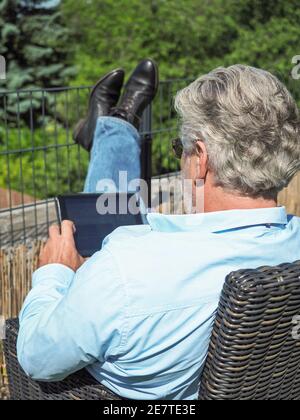 Senior man with grey hair sitting on a chair in the garden and looking to the tablet. From behind.  Stock Photo