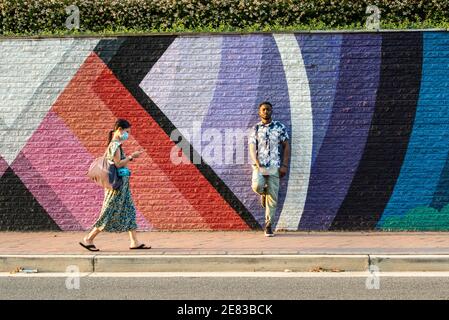 A Black man leans against a colorful street mural as a woman in a surgical mask walks by. Stock Photo