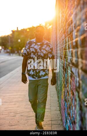 A Black man walks past a colorful mural in an urban setting as the sun goes down behind him. Stock Photo